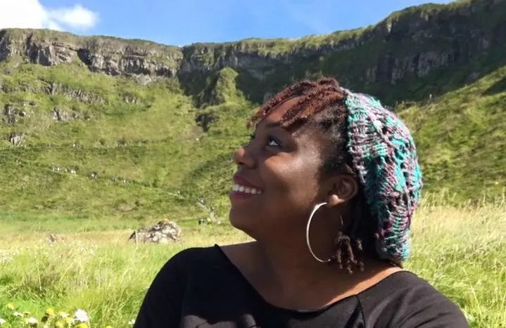 Meredith smiling to the left with a knitted cap on in a valley surrounded by green cliffs
